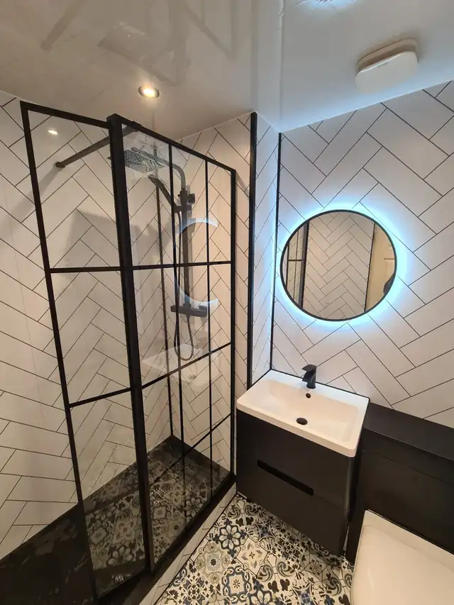 Walk-in shower with a black-framed glass screen, white herringbone wall tiles, a round backlit mirror and a black vanity unit