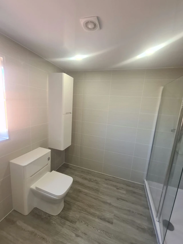 Bathroom with a white back-to-wall toilet, a tall white wall cabinet, beige wall tiles, a wood-effect floor and a glass shower screen at the side