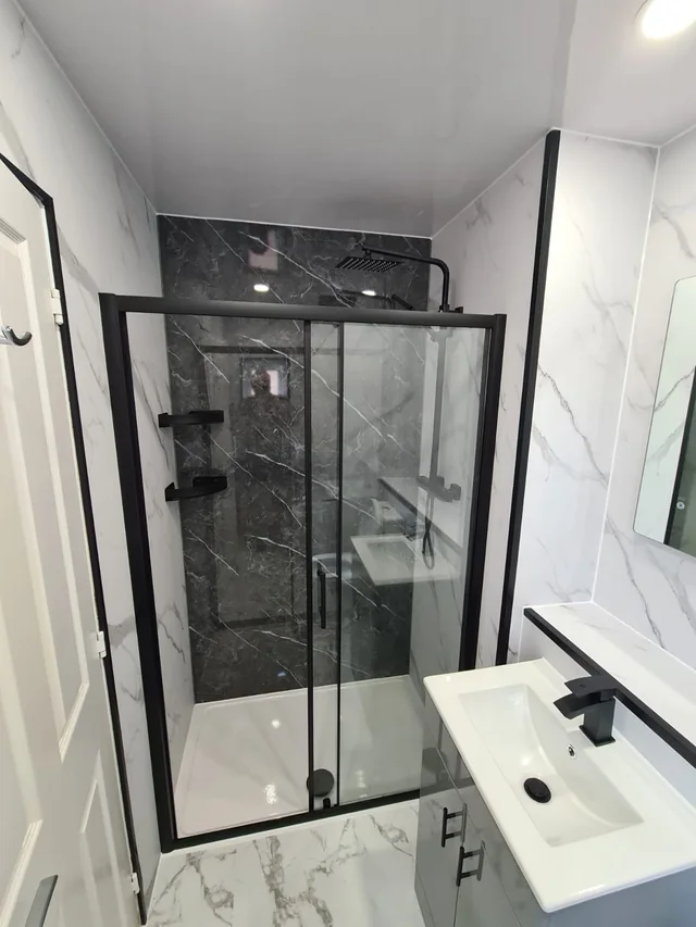 Shower enclosure with a black-framed sliding glass door, a dark marble-effect back wall, white marble-effect side walls and a white shower tray, with a basin vanity beside it