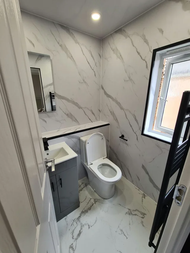 Small cloakroom with marble-effect wall panels and floor, a white toilet, a compact grey vanity with a black tap, a black-framed window and a black towel radiator
