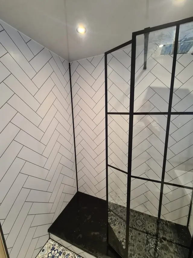 Corner shower with white herringbone wall tiles, a black-framed glass screen and a black shower tray, with patterned floor tiles outside