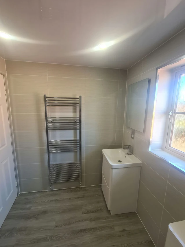 Bathroom with a tall chrome towel radiator, a white vanity basin with a chrome tap, a wall mirror, a window and beige wall tiles above a wood-effect floor