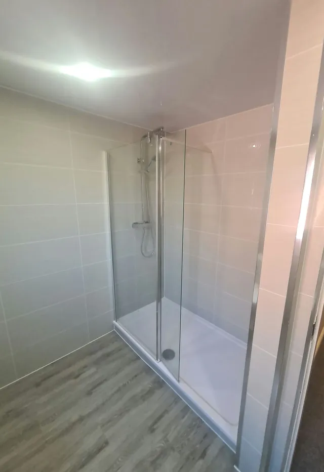 Walk-in shower with a glass screen, chrome thermostatic shower set and white shower tray, beside grey wall tiles and wood-effect flooring