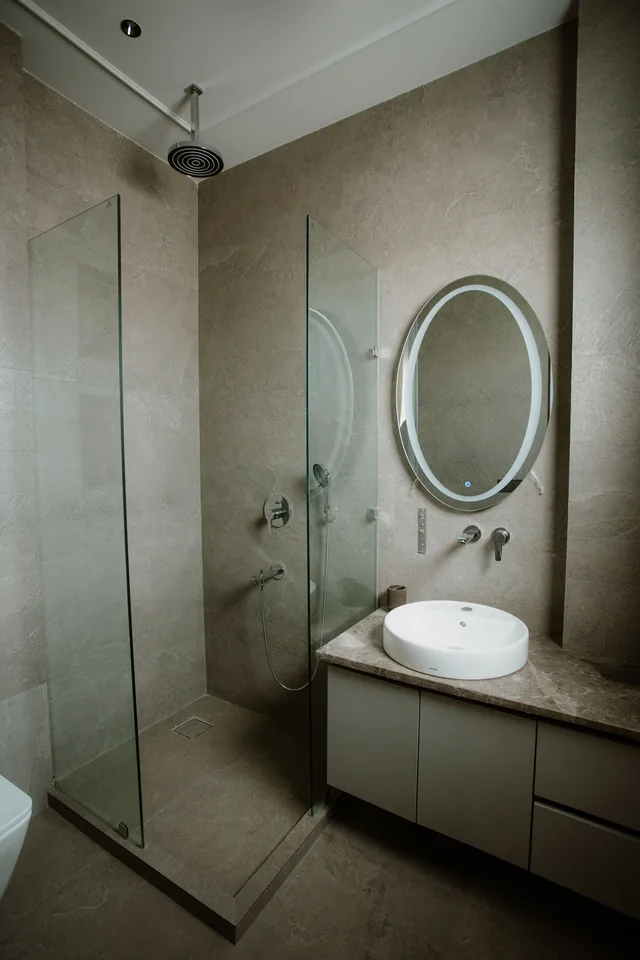Corner shower with two glass panels, a ceiling-mounted round shower head and stone-effect wall tiles, beside a wall-hung vanity with a round basin, an oval backlit mirror and wall-mounted taps