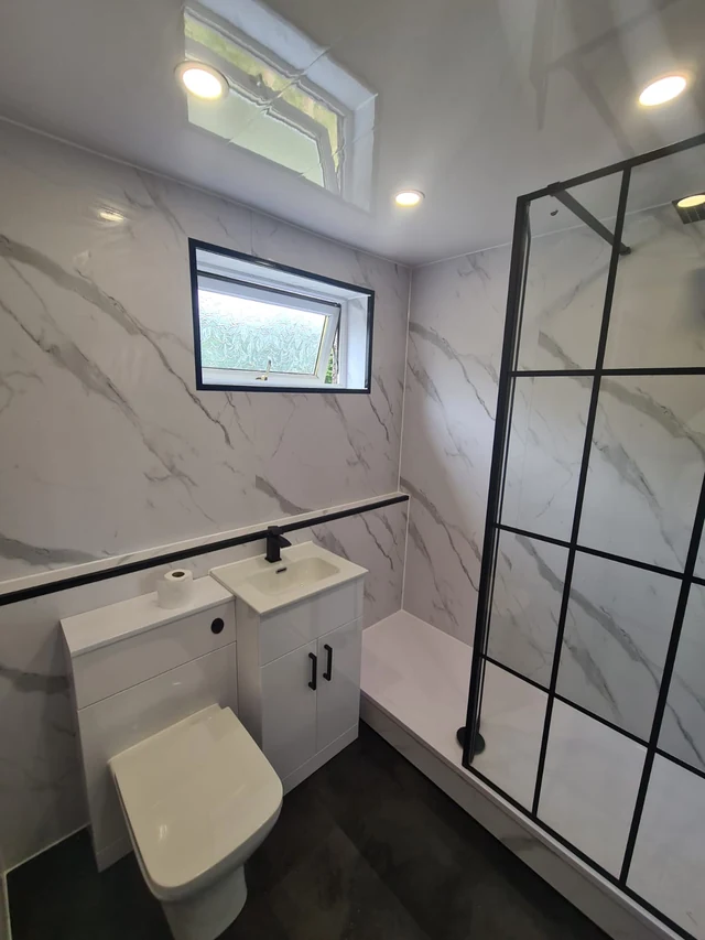 Bathroom with marble-effect wall panels, a black-framed glass shower screen, a white shower tray, a white toilet unit with a compact vanity and a small black-framed window