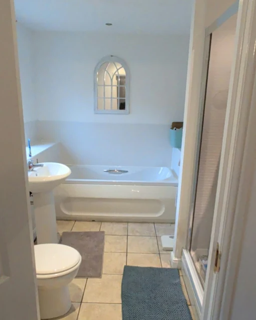 Bathroom before the refit, seen through a doorway: a white bath, pedestal basin and toilet, a tiled floor and a shower enclosure at the side